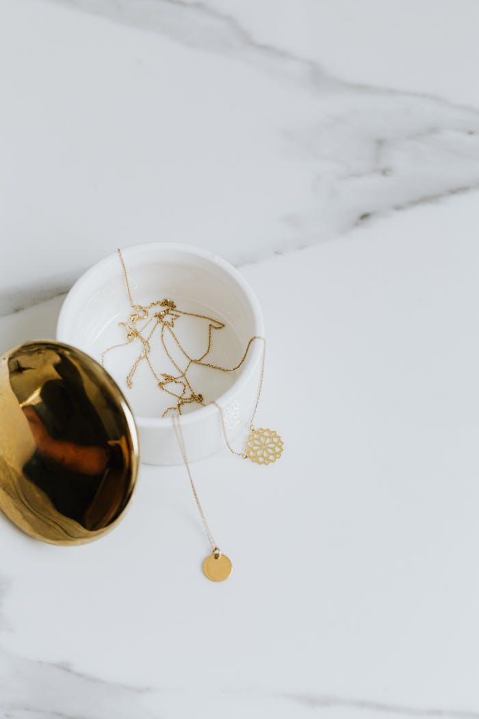 A minimalist photo of gold necklaces and pendants in a white marble container with a reflective lid.